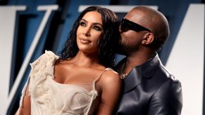 Kim Kardashian West is dressed in an ivory dress with Kanye West in a black shiny suit jacket in front of a blue, black and white background.