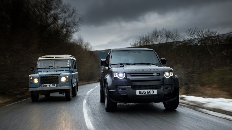 A 1977 Land Rover Defender Series III Stage One V8 and a 2021 Land Rover Defender 90 V8 travel side-by-side on a wet road