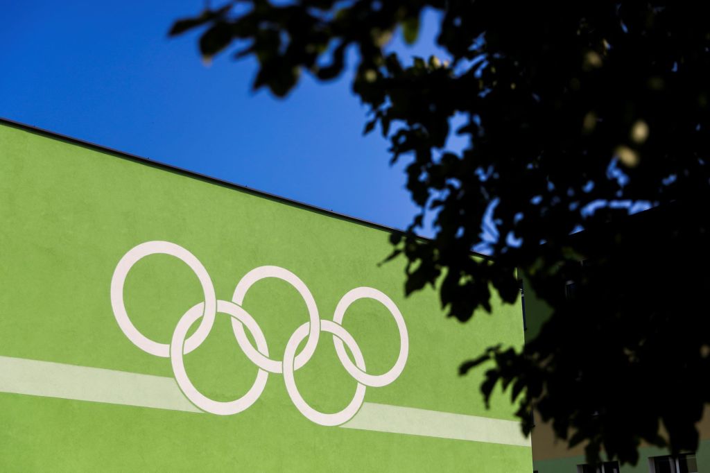 White Olympic rings painted on a green building with a blurred tree in the foreground to the left. 
