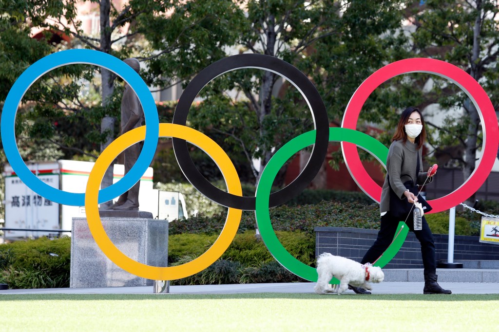 A woman wearing mask walks her pet dog past the 2020 Olympic rings
