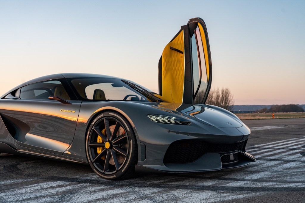 A grey 2021 Koenigsegg Gemera sits on a runway at sunset with the driver's door sticking straight up into the air 