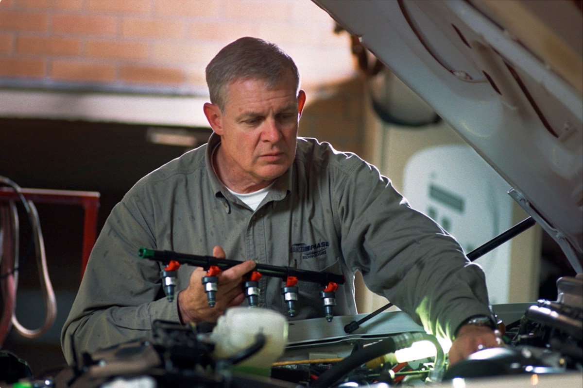 An automotive technician holding a fuel rail while under the hood of a car.
