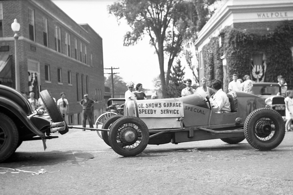 A black-and-white photo of an early racecar being towed by a rope through a town