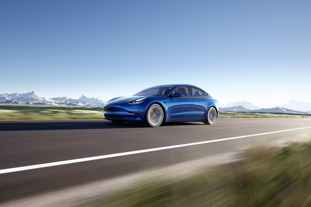 A blue Tesla Model 3 blasts it's way down a picturesque open road with mountains in the background