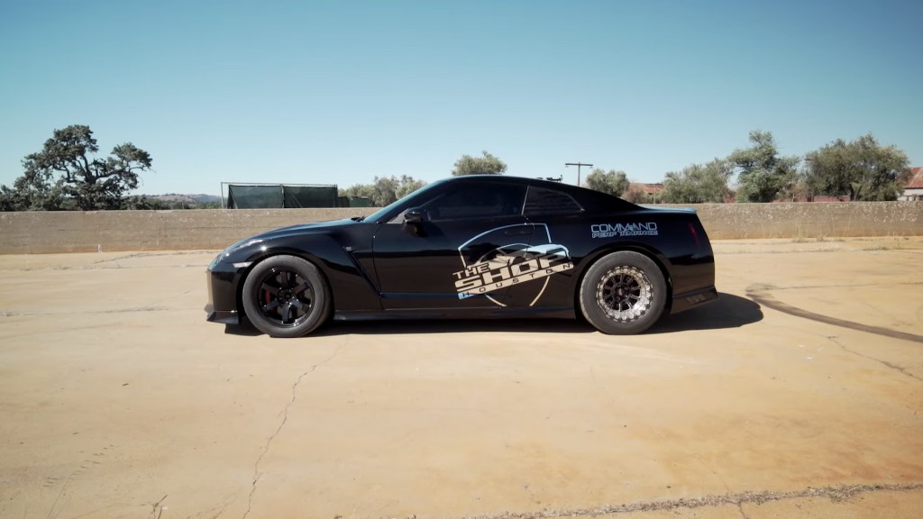 "Dancing Dan" Rue's 1,400 horsepower 2017 Nissan GT-R drag race car. The car is black with thick sidewall tires. There is a chrome graphic on the side of the car that reads "The SHOP Houston"