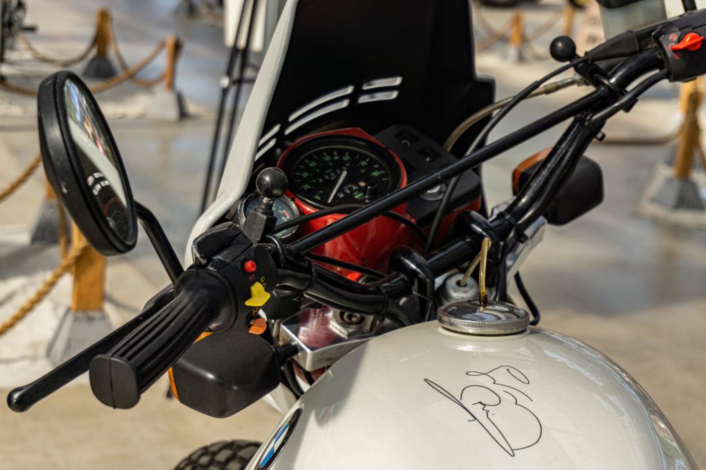 A close-up view of a white 1987 BMW R80 G/S's handlebars, autographed fuel tank, and red-trimmed gauges