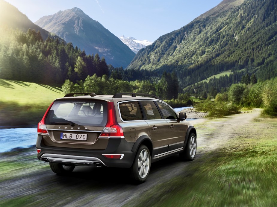 A bronze 2014 Volvo XC70 wagon traveling on a dirt road along a river through a mountain valley