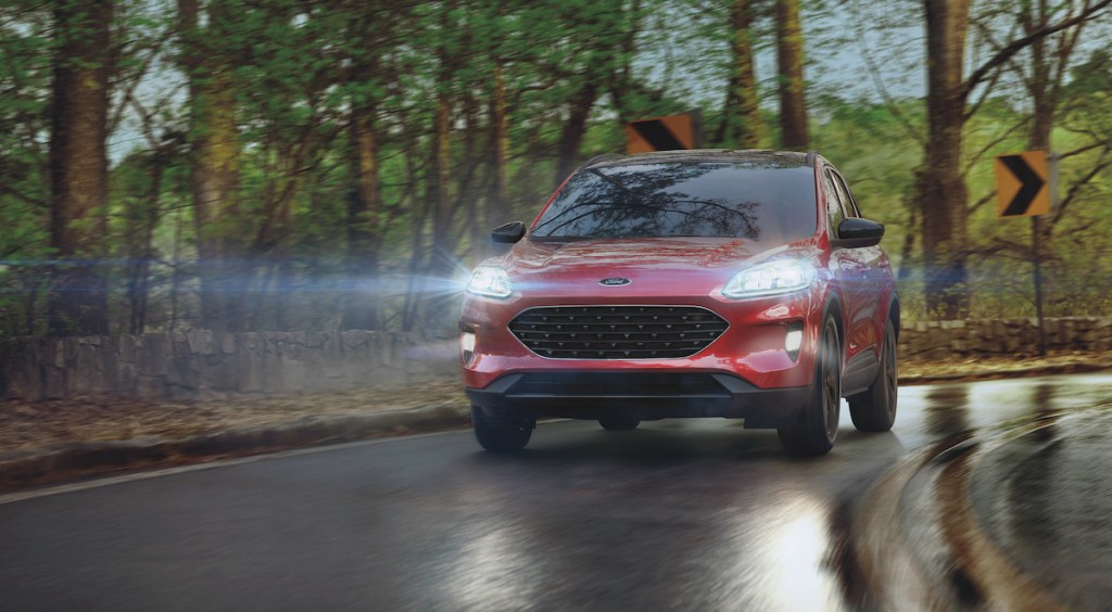 A red 2021 Ford Escape compact SUV with its headlights illuminated travels on a wet road past trees on a rainy day