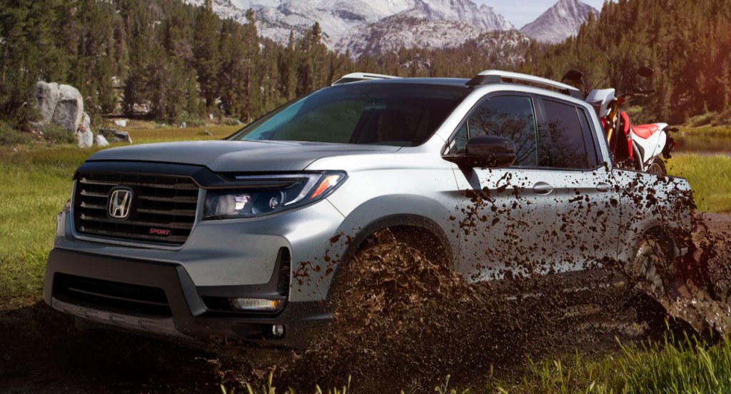 A silver Honda Ridgeline mid-size truck driving through mud while carrying a motorbike in its truck bed.