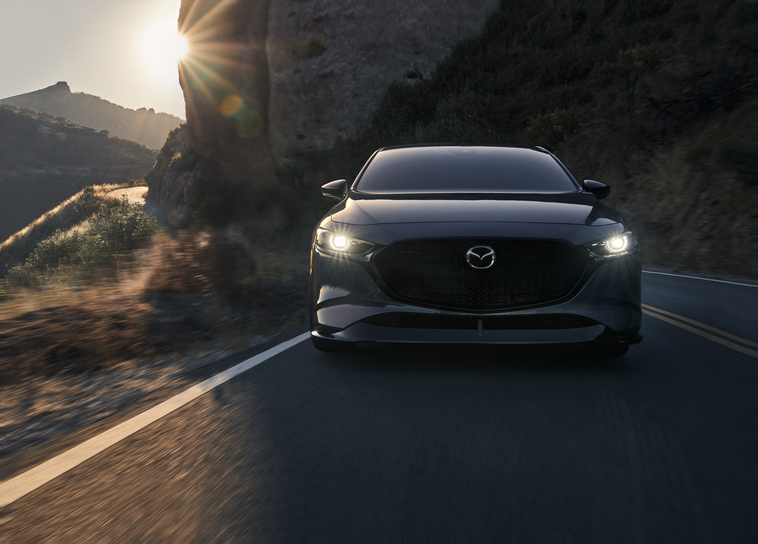 A grey Mazda 3 AWD hatchback shot from the front on a canyon road at sunset