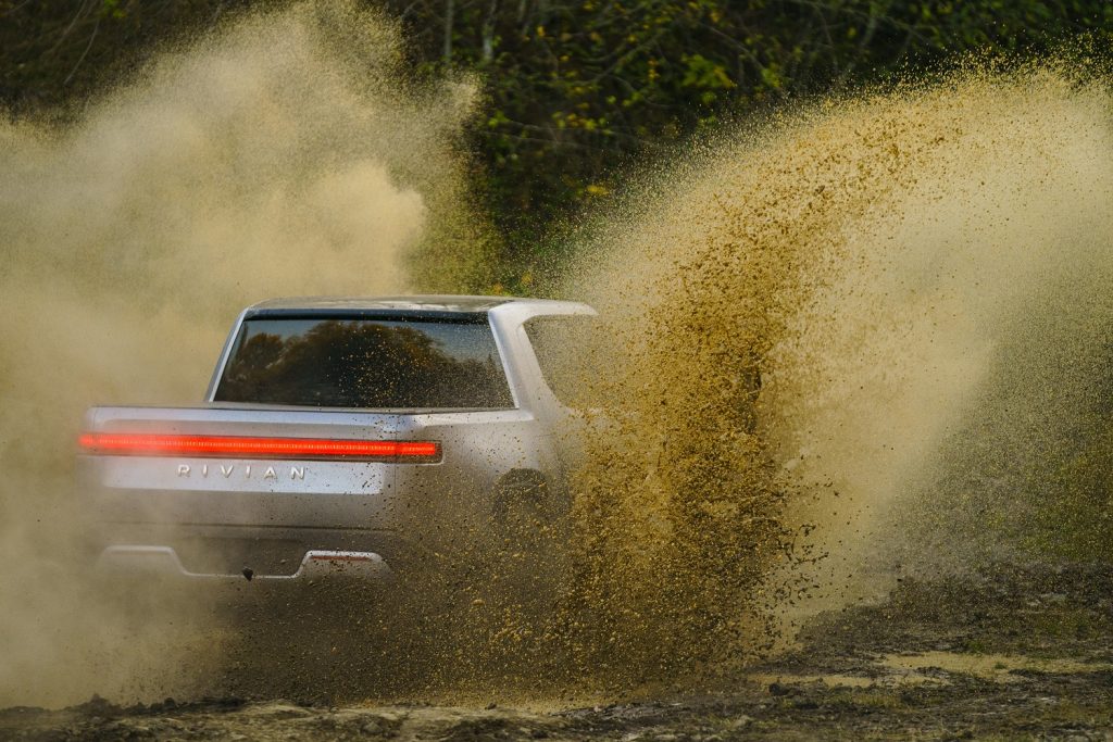 This is a press photo of a silver R1T Rivian electric truck driving through dirt. Motor Trend reviewed the capabilities of the Rivian off road