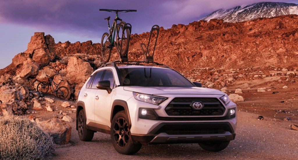 A white 2021 Toyota Rav4 TRD Off-Road is on a trail with mountains in the background. The SUV has a bike roof rack with a bike fastened to it. 