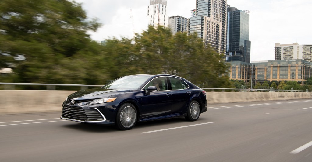 A new 2022 Toyota Camry rolls down a city highway with dark blue paint and a softer front end than last year's model