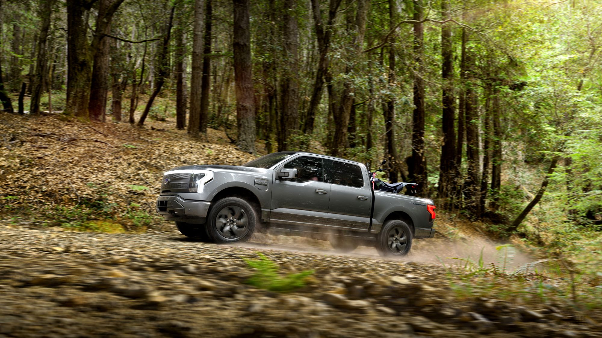 A gray 2022 Ford F-150 Lightning driving on a dirt road in a wooded area.