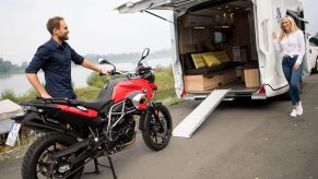 A man loads a red BMW motorcycle onto a white Knaus camper van RV with a ramp