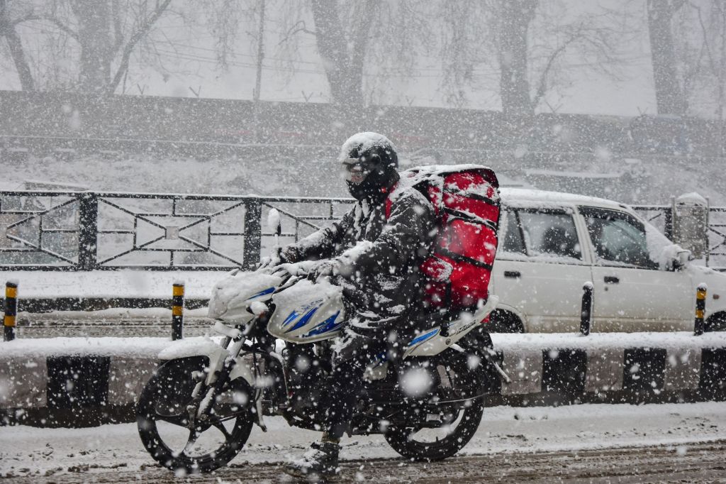 A black-clad motorcycle rider dressed in full winter gear rides through a Kashmir snowstorm