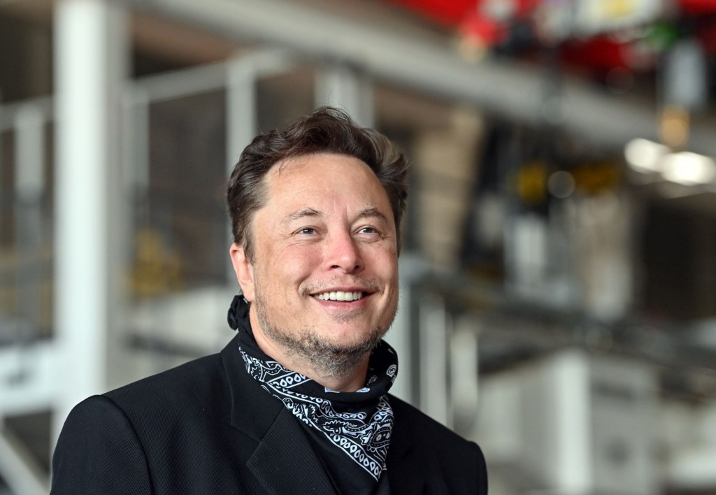 Elon Musk dressed all in black with a black and white handkerchief around his neck with industrial pipes in the background. 