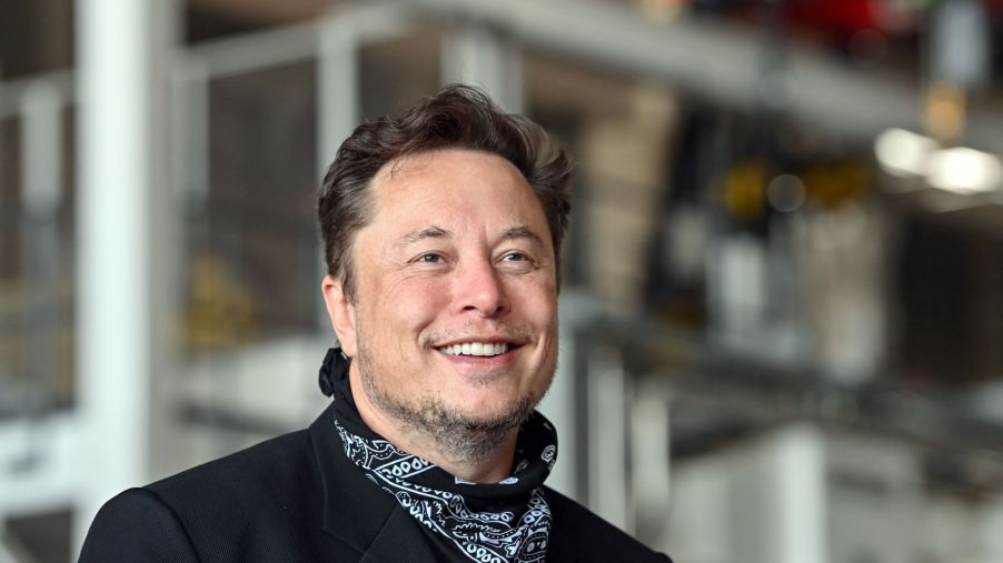 Elon Musk dressed all in black with a black and white handkerchief around his neck with industrial pipes in the background.