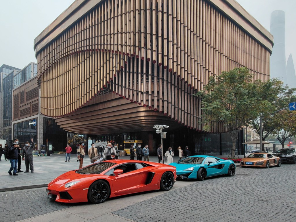A red Ferrari and a light blue Lamborghini in front of a modern shaped tall building.