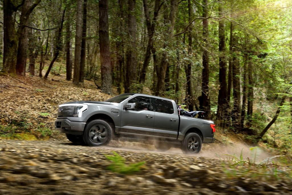 Gray 2022 Ford F-150 Lightning driving through a forest with a motorbike in its truck bed. 
