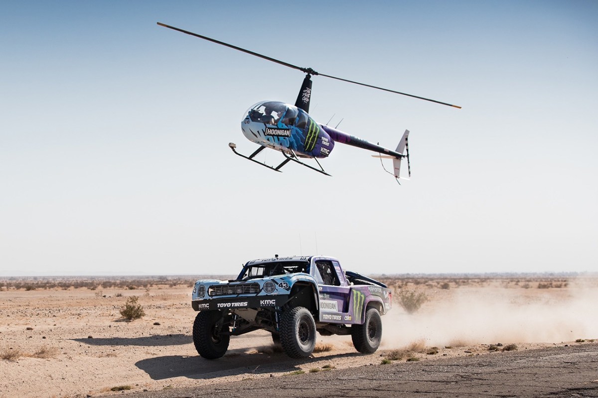 Hoonigan race truck driving through the desert with a Hoonigan helicopter overhead.