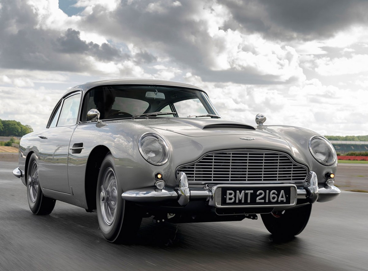 A sliver Aston Martin DB5 James Bond continuation model driving on a race track with a cloudy sky in the background.