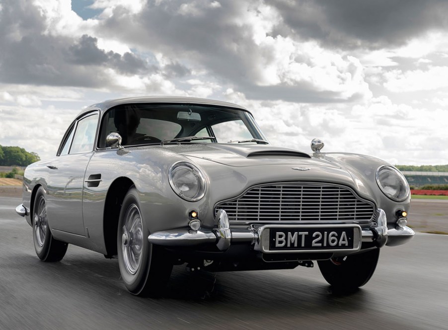 A sliver Aston Martin DB5 James Bond continuation model driving on a race track with a cloudy sky in the background.