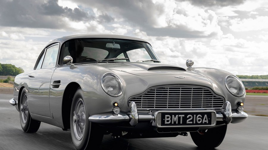 A sliver Aston Martin DB5 James Bond continuation model driving on a race track with a cloudy sky in the background.