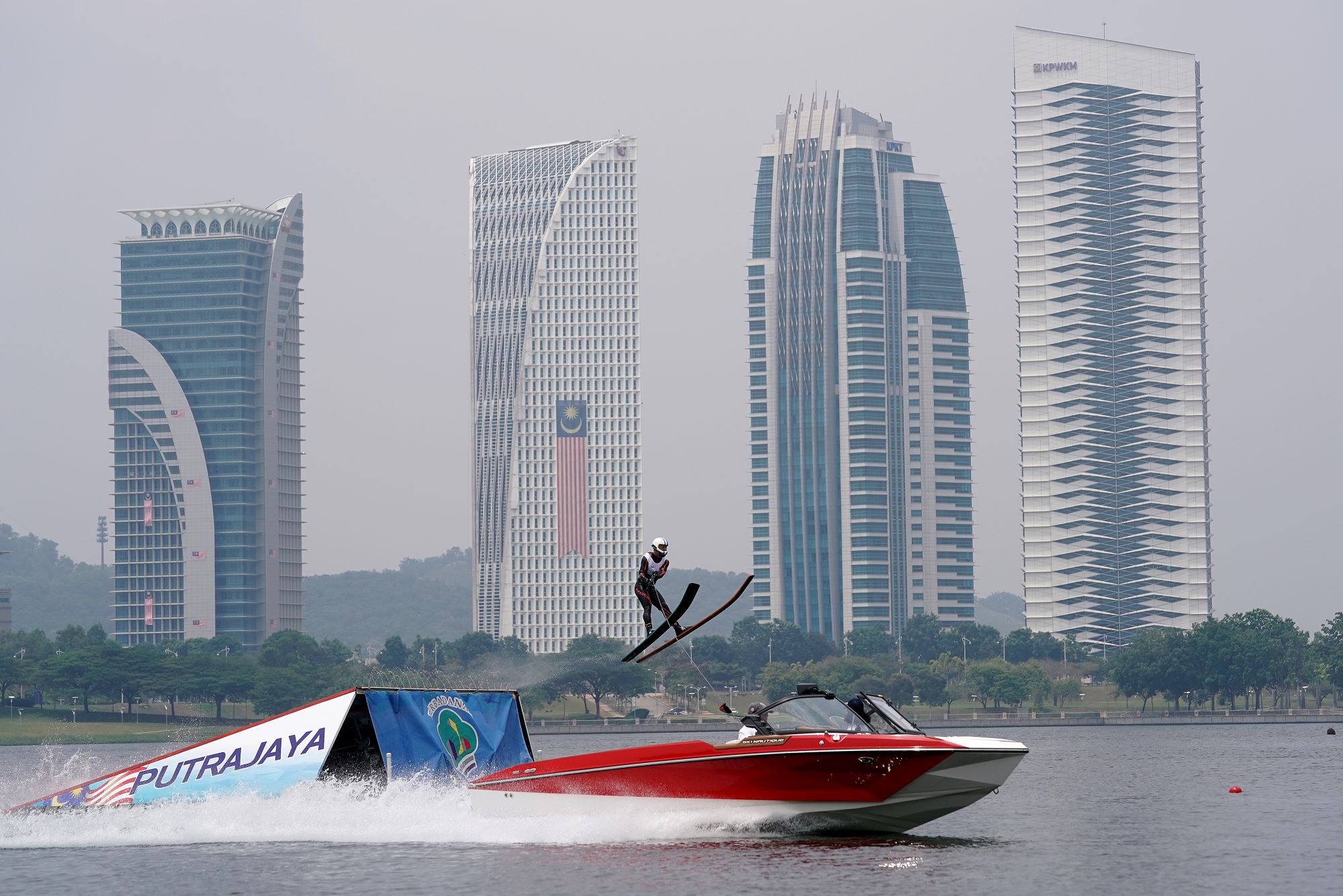 Jet boat on the water with a someone riding skis going up a ramp in front of a city skyline.