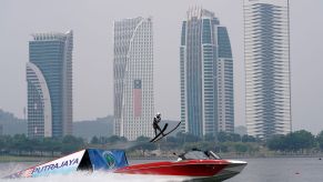 Jet boat on the water with a someone riding skis going up a ramp in front of a city skyline.