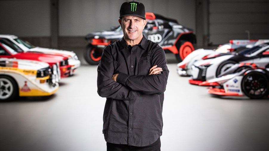 Rally racer Ken Block standing in a room with Audi rally cars and other race cars behind him