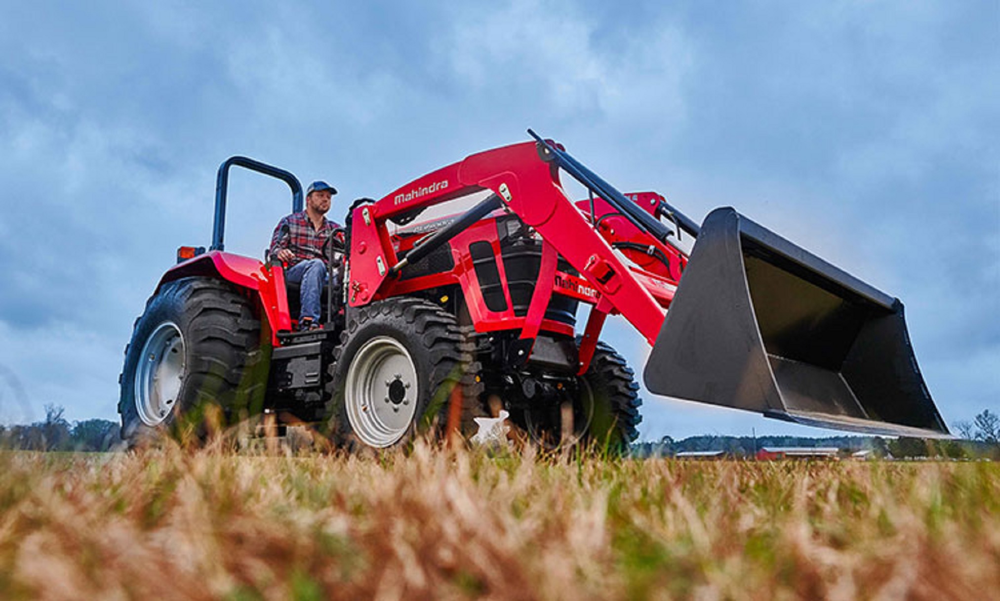 A farmer operates a red Mahindra 5100 Series utility tractor in a field