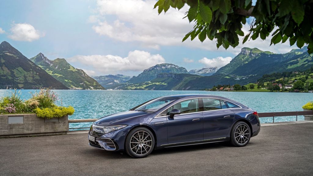 A blue Mercedes-Benz EQS sitting on a concrete area in front of a large body of water with mountains in the background.
