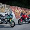A green-and-silver and a red Moto Guzzi V100 Mandello on rear-wheel stands by a graffiti-covered brick wall