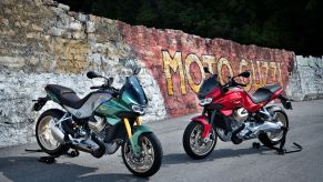 A green-and-silver and a red Moto Guzzi V100 Mandello on rear-wheel stands by a graffiti-covered brick wall