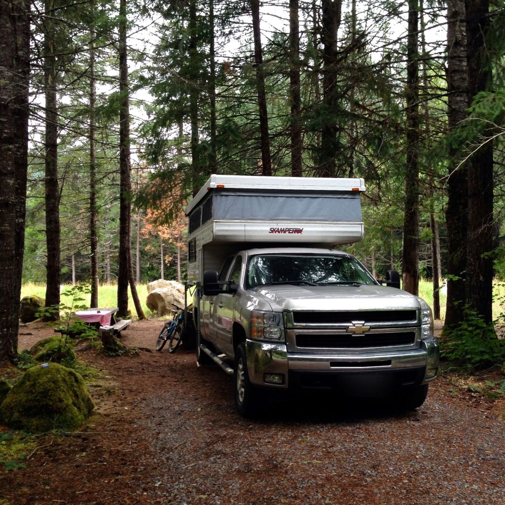 A silver truck pulling an off-road camper in a wooded camping area.