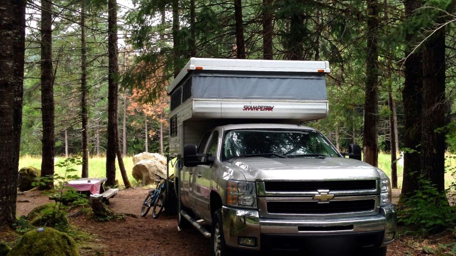 A silver truck pulling an off-road camper in a wooded camping area.