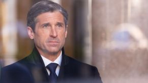 Patrick Dempsey looking through a window with a few reflections form the outside dressed in a black suit.