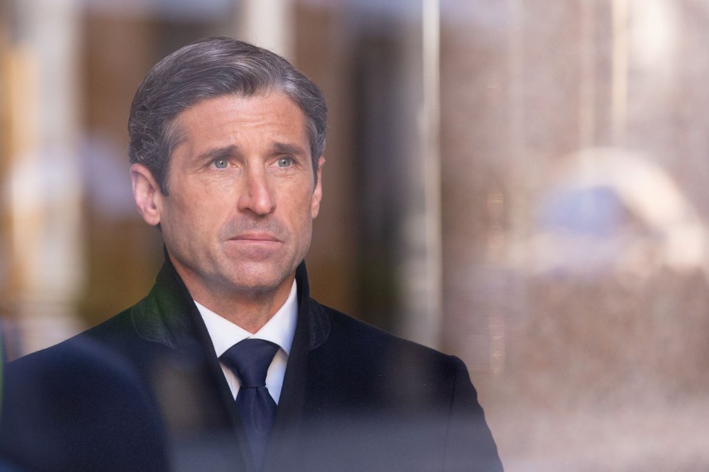 Patrick Dempsey looking through a window with a few reflections form the outside dressed in a black suit.