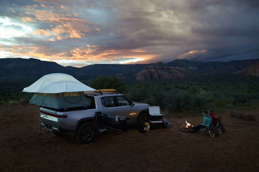 This is a press photo of a silver R1T Rivian electric truck outfitted with a rooftop tent in the mountains. Motor Trend reviewed the capabilities of the Rivian off road