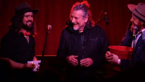 Robert Plant dressed in black on stage with a red curtain in the background.