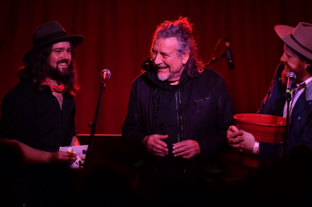 Robert Plant dressed in black on stage with a red curtain in the background.