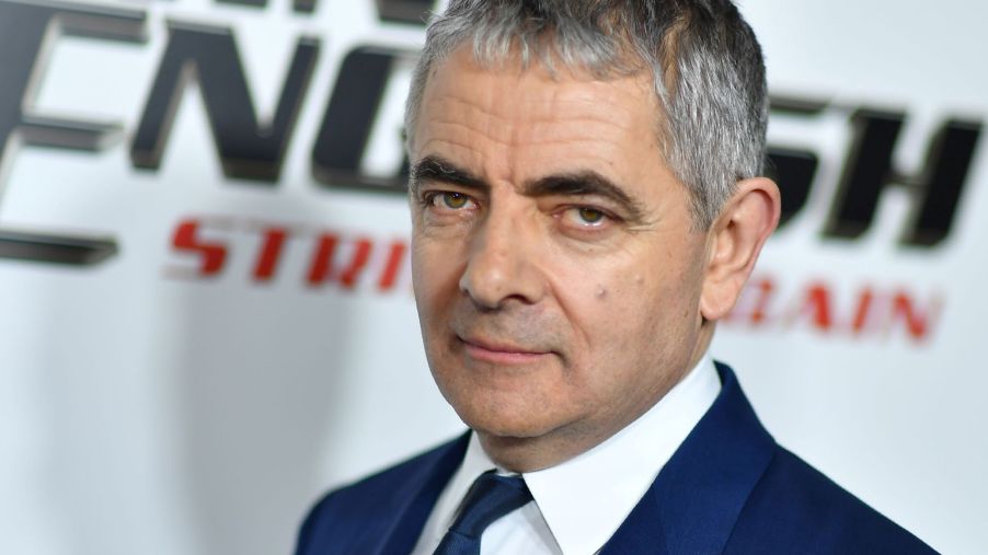 Rowan Atkinson dressed in a blue suit standing in front of a white background with black and red writing.