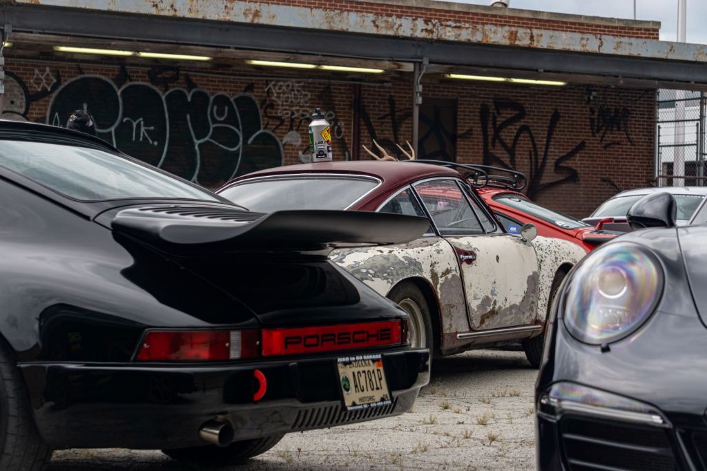 Several classic Porsche 911s with a modern Panamera in a parking lot next to a graffiti-sprayed brick wall