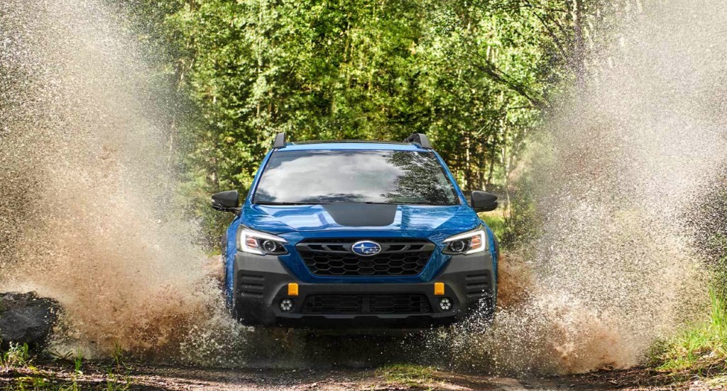 A blue Subaru Outback Wilderness is off-roading in the mud surrounded by trees with green leaves.  