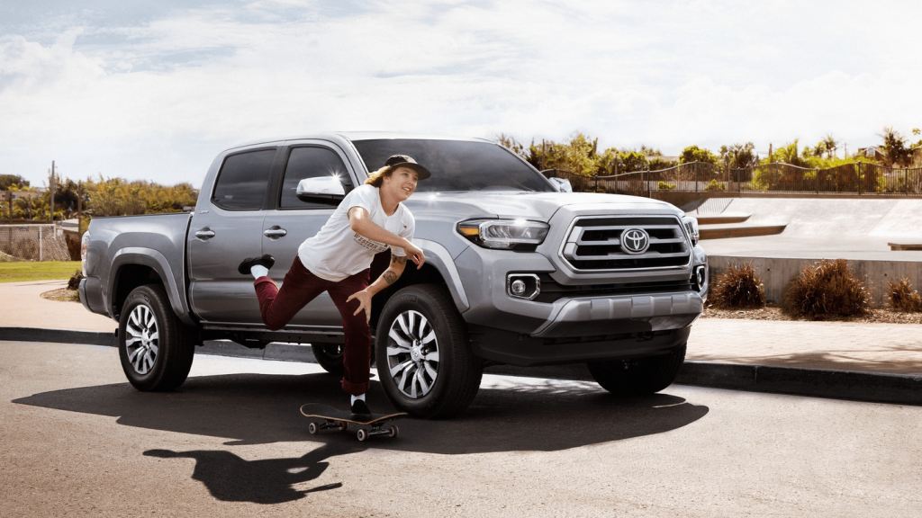 A silver Toyota Tacoma parked outside with a skateboarder going by. 