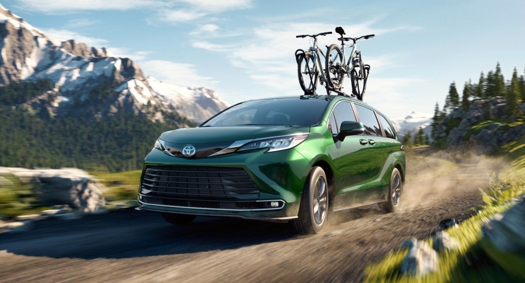 A green 2022 Toyota Sienna minivan is driving down a gravel road with two bikes attached to its roof. 