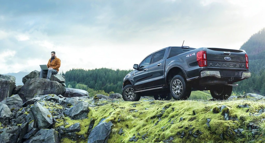 A black Ford Ranger compact pickup truck is parked on a mountain. A man is on his laptop sitting on a rock nearby. 