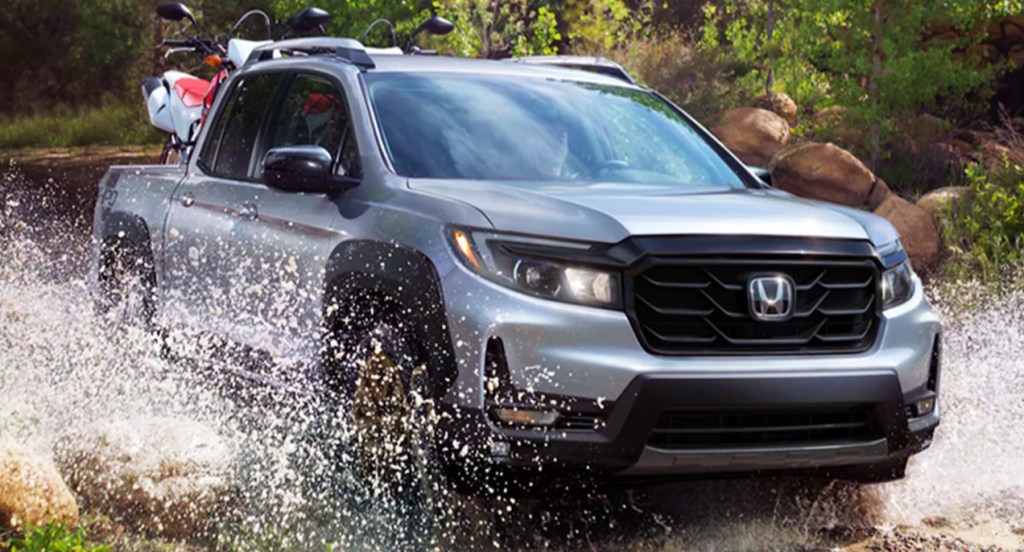 A silver Honda Ridgeline crosses a small, shallow body of water while carrying two dirt bikes in its truck bed. The Honda Ridgeline truck appears to be in the wilderness. 