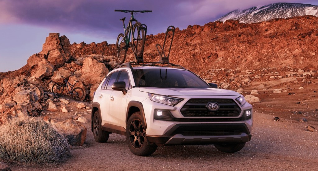 A white Toyota RAV4 TRD Off-Road SUV is parked in a mountainous region. One bike is loaded atop the SUV. Another bike is in the background. 